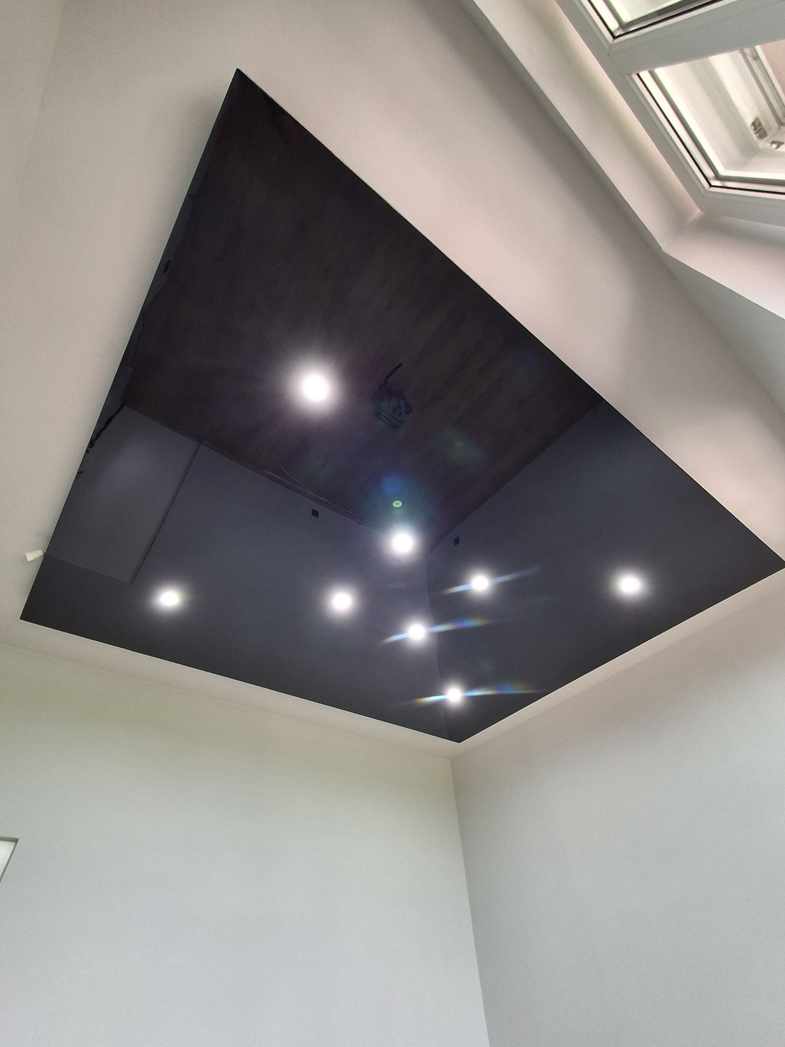 Błyszczący, czarny sufit z wbudowanymi punktami świetlnymi, odbijający światło, w rogu widoczne okno dachowe, białe ściany. Nowoczesne wykończenie wnętrza.