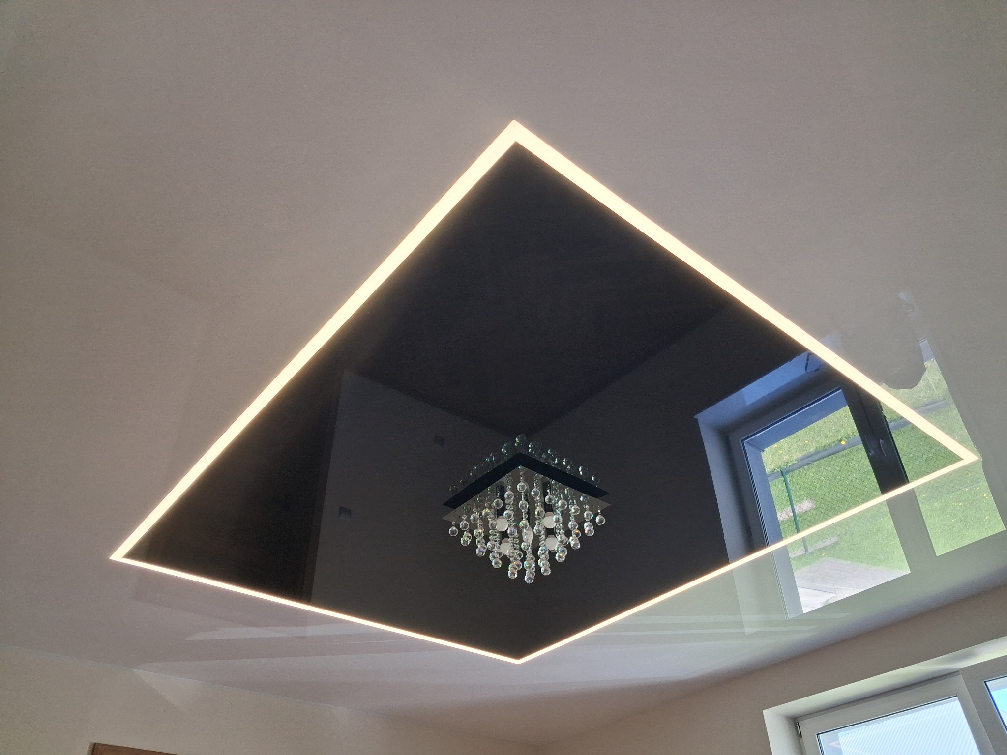 Błyszcząca, czarna powierzchnia sufitu z kwadratowym podświetleniem LED, odbijająca kryształowy żyrandol i okno z widokiem na zieleń. Nowoczesny design.