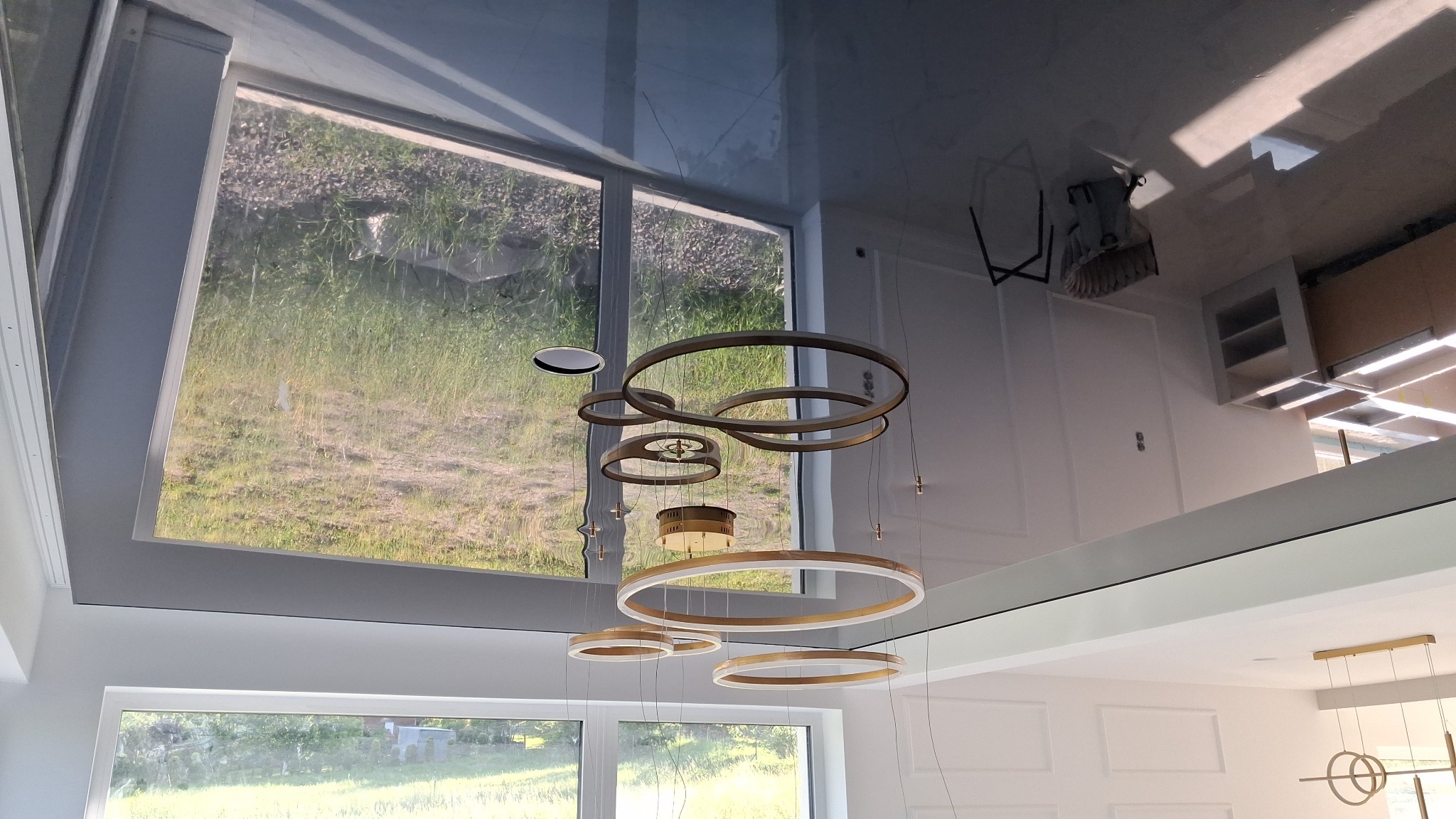 Błyszczący, czarny sufit napinany odbija nowoczesny żyrandol i okna, tworząc iluzję przestrzeni w jasnym, minimalistycznym wnętrzu.