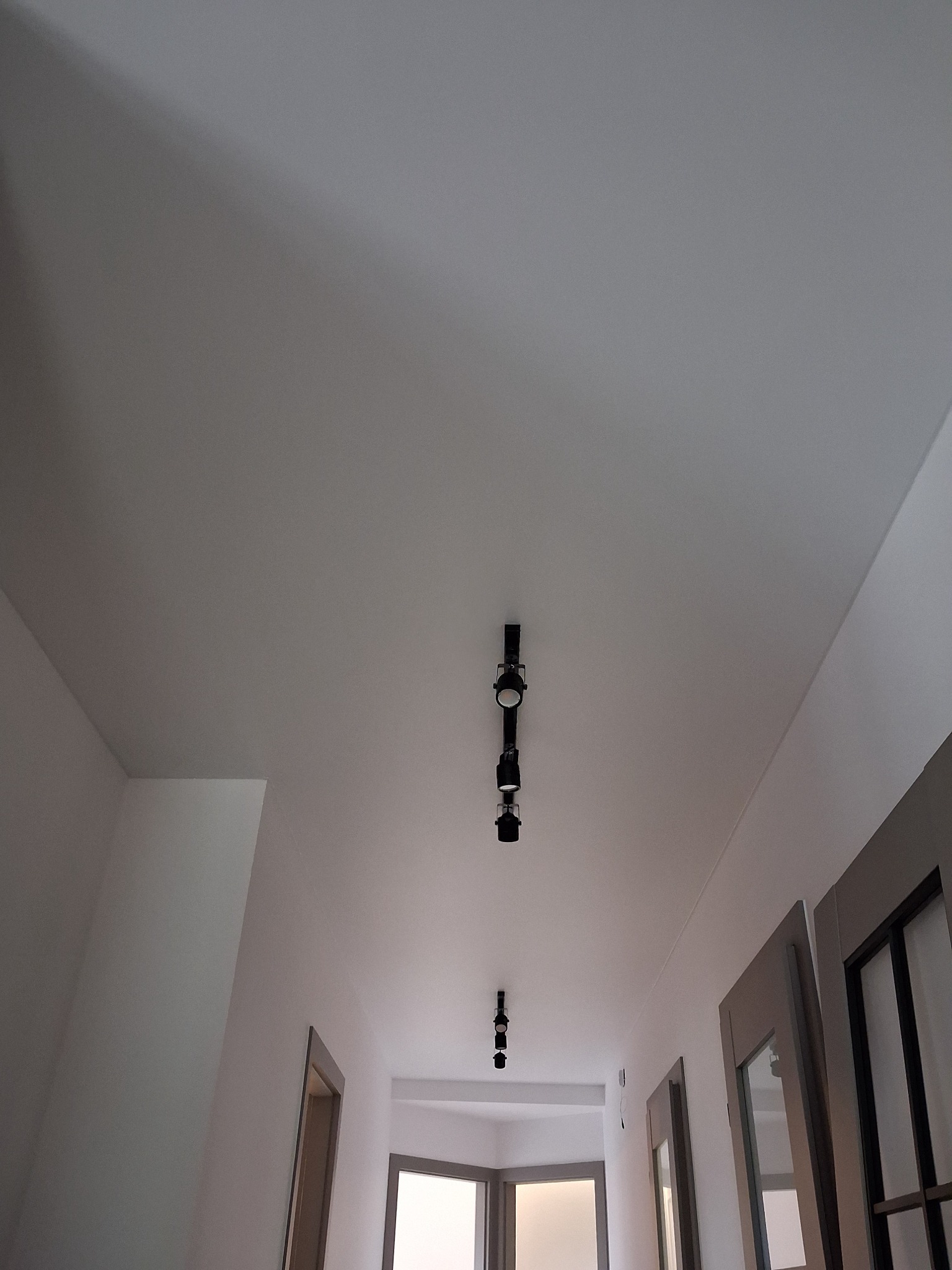 Biały sufit z zamontowanym czarnym oświetleniem punktowym na szynie, widok z dołu w korytarzu. Widoczne drzwi i fragment okna w oddali.