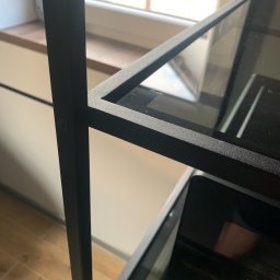 Prokonekt-met - Metalowa konstrukcja regału z półkami z ciemnego szkła, widoczny detal łączenia elementów na śrubę, w tle okno.