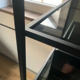 Metalowa konstrukcja regału z półkami z ciemnego szkła, widoczny detal łączenia elementów na śrubę, w tle okno.