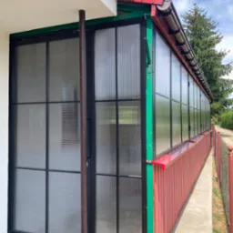 Zadaszona weranda z czarnymi metalowymi ramami okiennymi i drzwiami, wypełniona półprzezroczystym, prążkowanym tworzywem sztucznym, z czerwonymi elementami wykończeniowymi i zielonymi narożnikami...