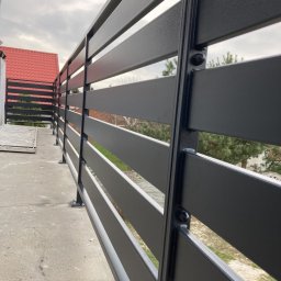 Prokonekt-met - Nowoczesne, poziome, metalowe ogrodzenie balkonowe w kolorze grafitowym, mocowane na śruby, z widokiem na czerwony dach budynku i drzewa w tle.