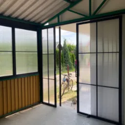 Zadaszona weranda z przesuwnymi, przeszklonymi drzwiami z czarną ramą i ścianami z blachy trapezowej, rower zaparkowany na zewnątrz.