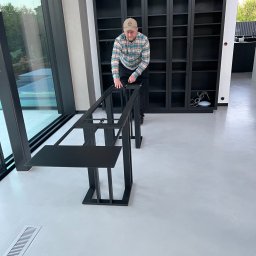 Prokonekt-met - Mężczyzna w czapce i kraciastej koszuli montuje czarną, metalową konstrukcję stołu w nowoczesnym wnętrzu z dużymi oknami i zabudowaną biblioteczką.