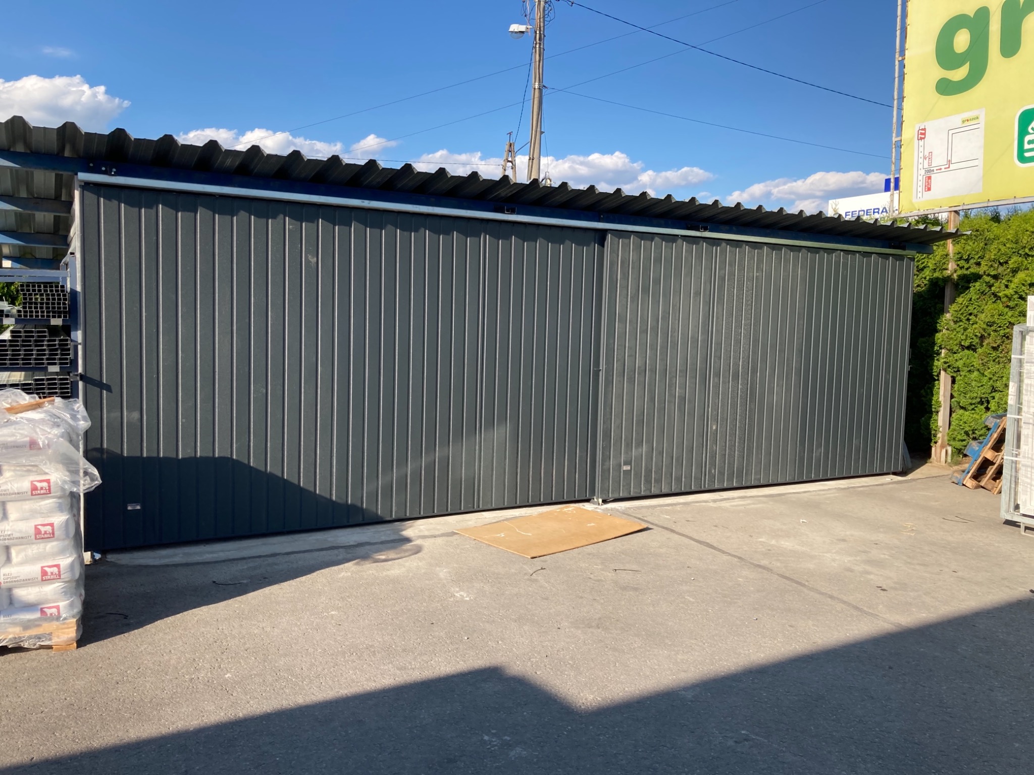 Szara hala z blachy falistej z przesuwnymi drzwiami, widok z zewnątrz, obok paleta z workami cementu.