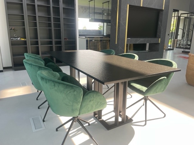 Stół z blatem o ciemnej, ziarnistej fakturze i metalową podstawą z pionowych prętów, otoczony zielonymi, welurowymi krzesłami na metalowych nogach. W tle widoczny regał na książki i fragment ściany...