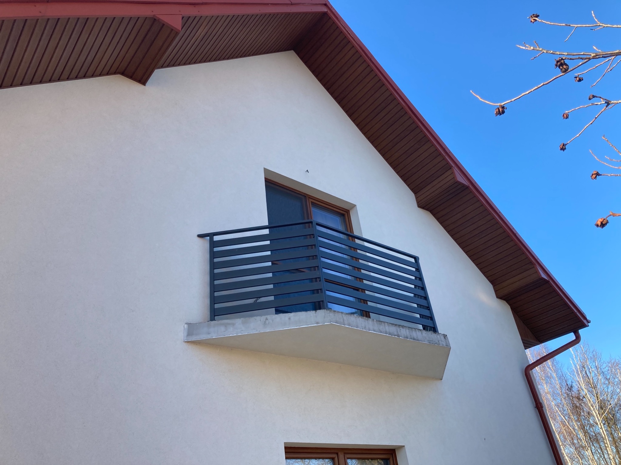 Elewacja domu z balkonem z metalową, poziomą balustradą pomalowaną na ciemny kolor, widok z dołu na tle błękitnego nieba i gałęzi drzew.