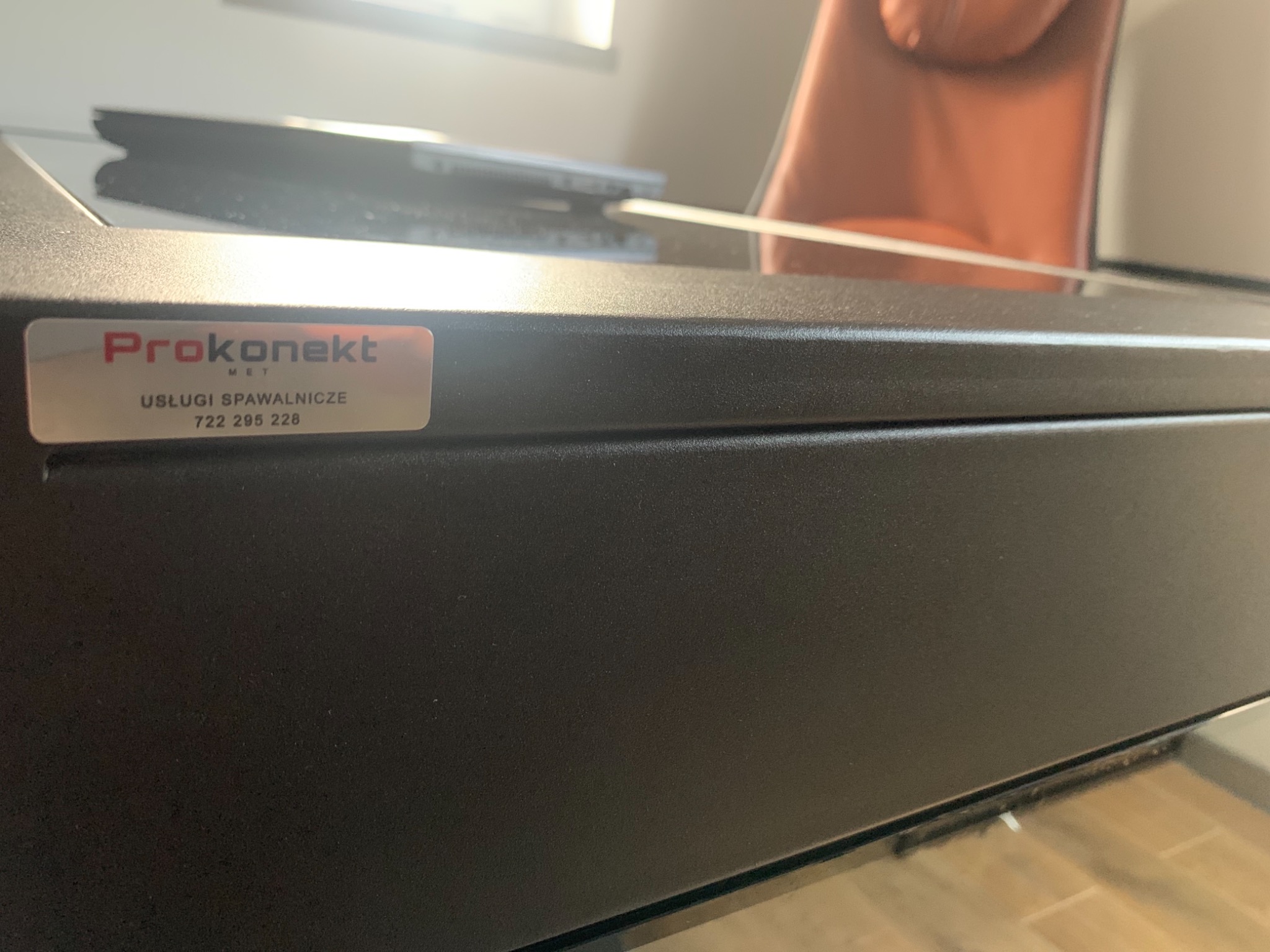 Ciemny, metalowy element konstrukcyjny z naklejoną etykietą informującą o usługach spawalniczych firmy Prokonekt MET, zawierającą numer telefonu.