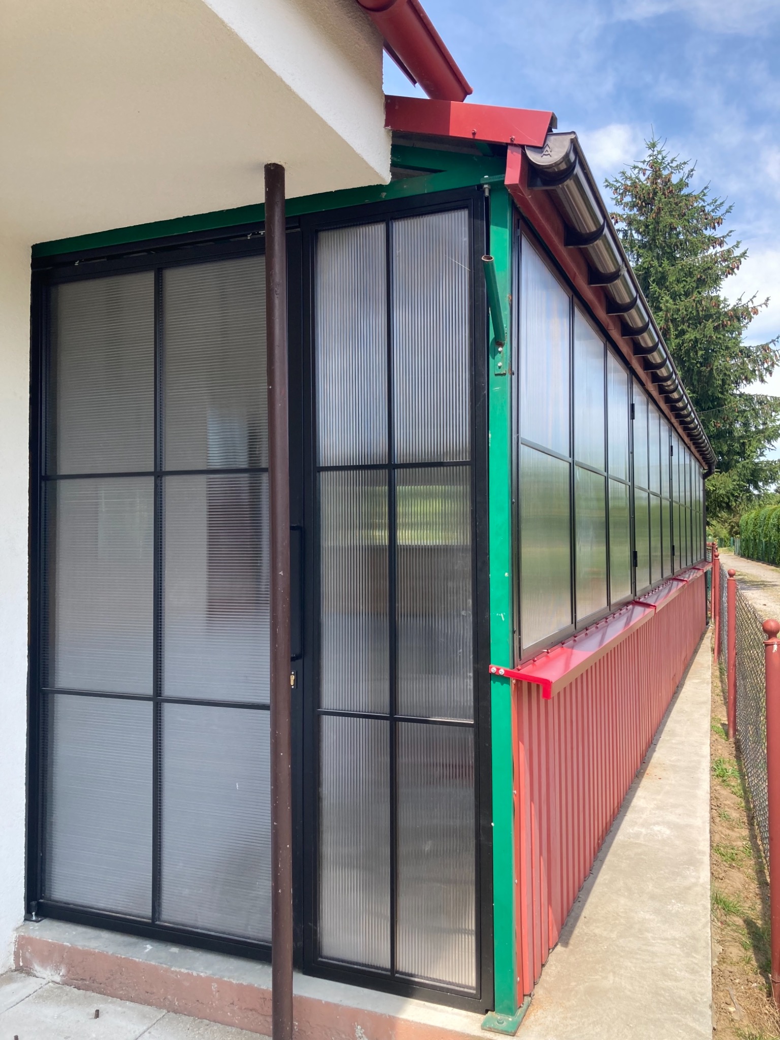 Zadaszona weranda z czarnymi metalowymi ramami okiennymi i drzwiami, wypełniona półprzezroczystym, prążkowanym tworzywem sztucznym, z czerwonymi elementami wykończeniowymi i zielonymi narożnikami...