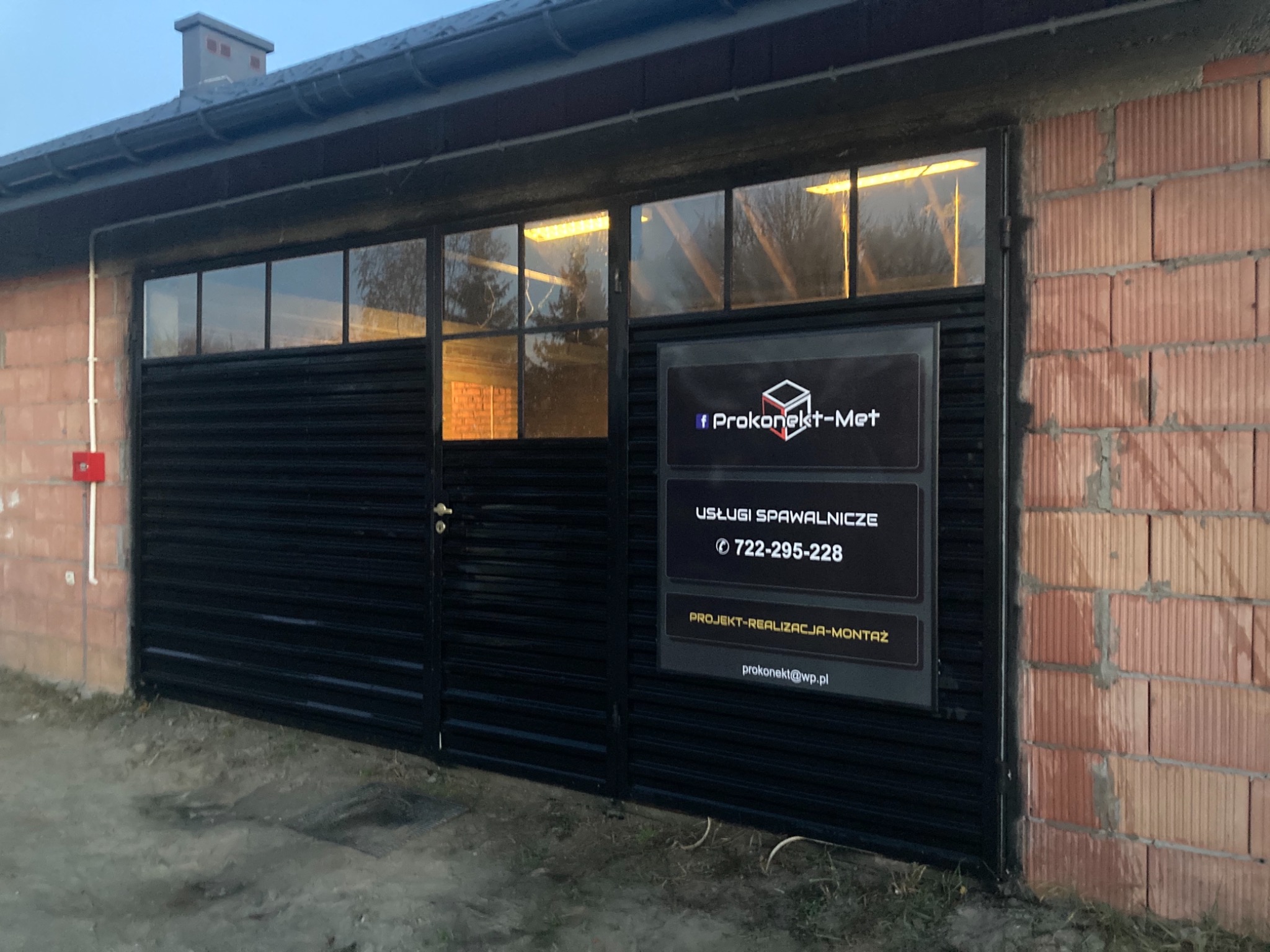 Czarna brama wjazdowa z poziomych profili zamontowana w budynku z cegły dziurawki, z tabliczką reklamową firmy Prokonekt-Met oferującej usługi spawalnicze.