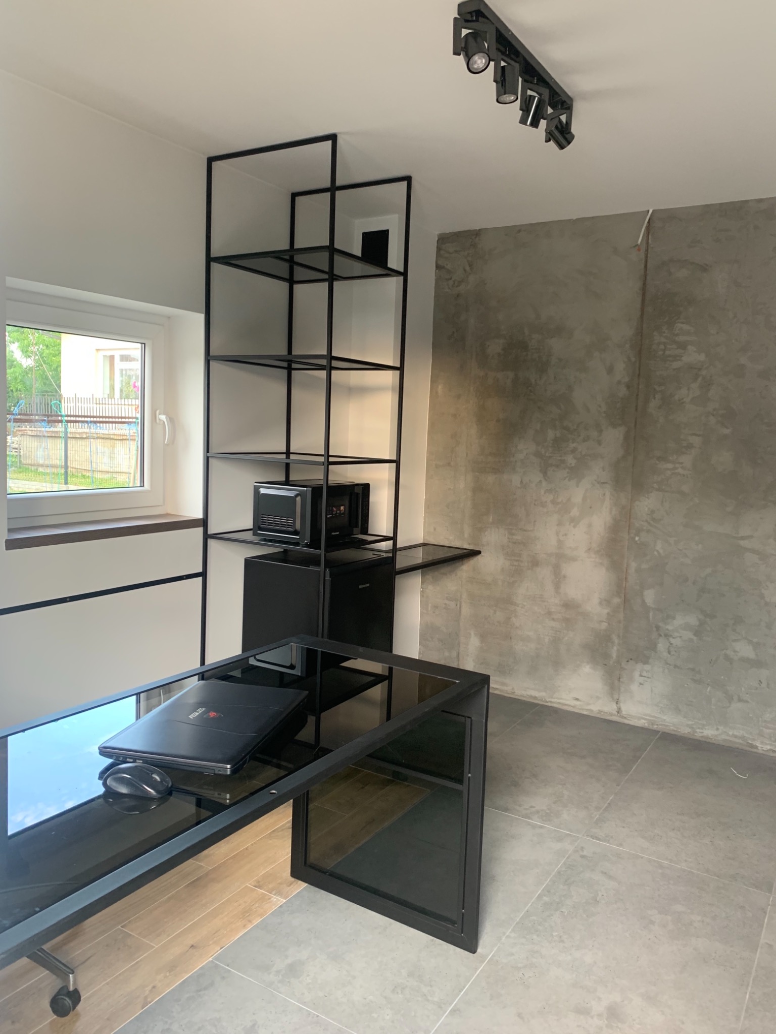 Nowoczesne biuro z czarnym metalowym regałem, szklanym biurkiem z laptopem i myszką, betonową ścianą w tle oraz oświetleniem punktowym na suficie.