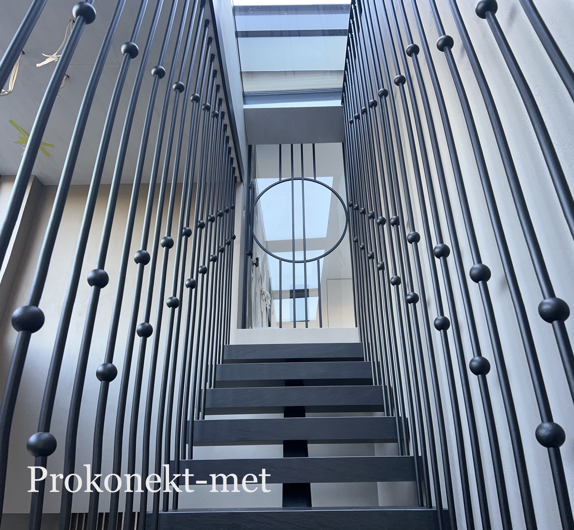 Nowoczesne schody metalowe z czarnymi poręczami zdobionymi kulami, prowadzące do przeszklonego dachu, widziane z dołu.