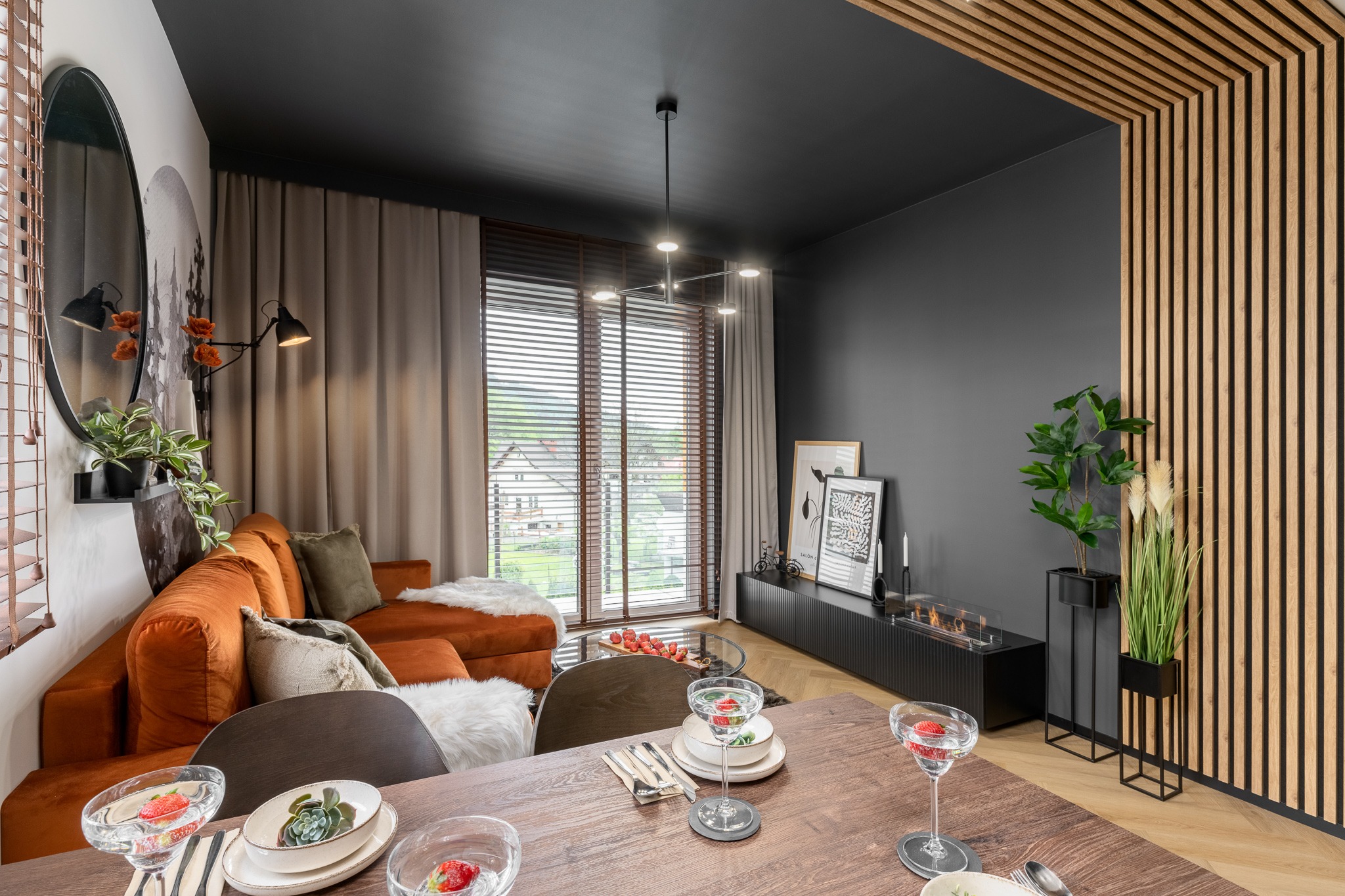 Nowoczesne wnętrze salonu z pomarańczową sofą, drewnianym stołem z nakryciem, czarnym sufitem i ścianą, dekoracyjną ścianką z drewnianych listew oraz biokominkiem.