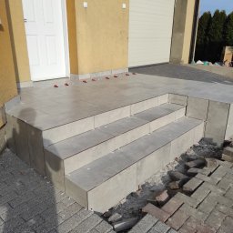 Mariusz Lasota - Wykończone płytkami schody wejściowe do domu, widoczne małe krzyżyki dystansowe, kostka brukowa częściowo usunięta, żółta elewacja budynku i białe drzwi.