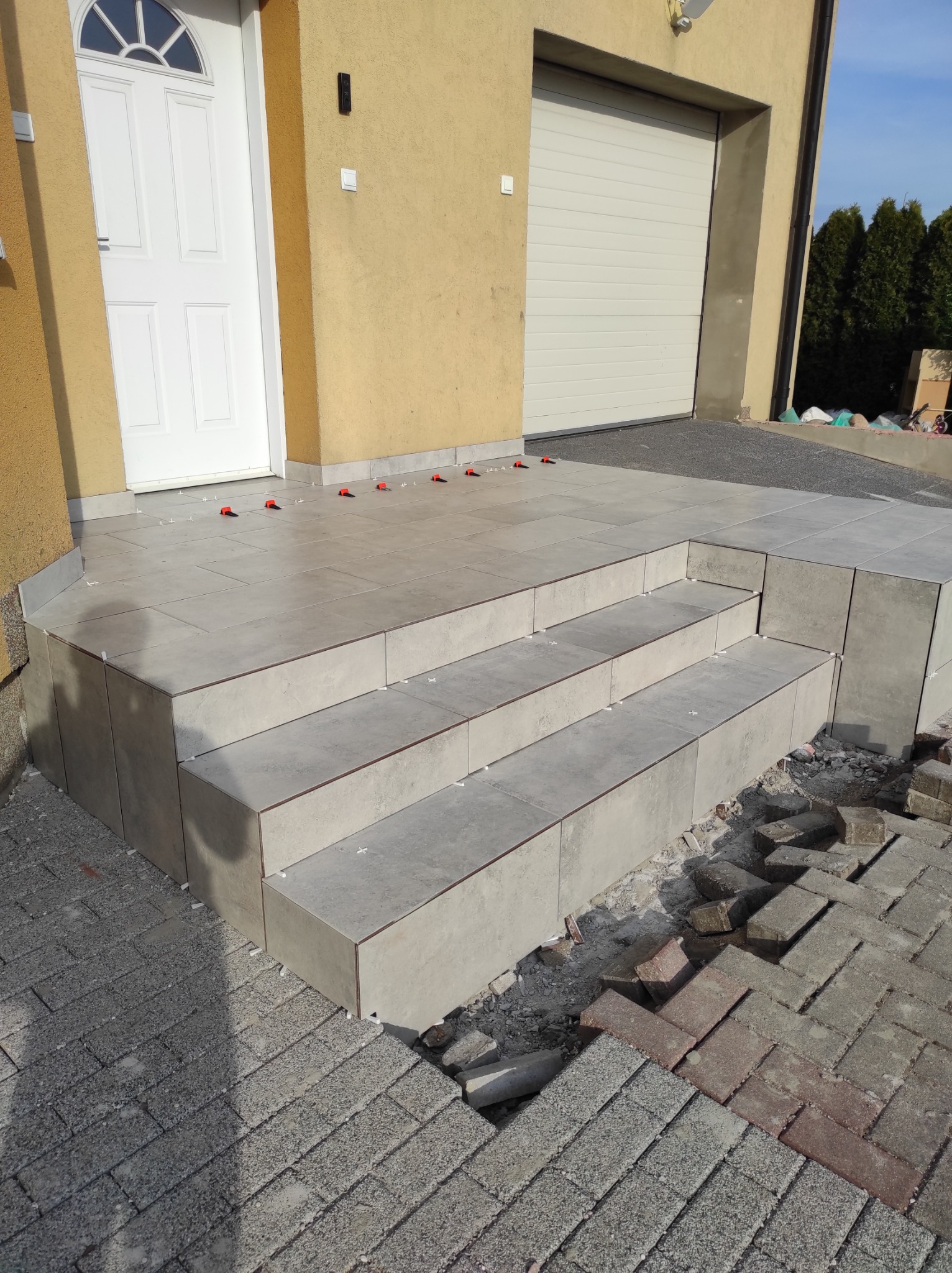 Wykończone płytkami schody wejściowe do domu, widoczne małe krzyżyki dystansowe, kostka brukowa częściowo usunięta, żółta elewacja budynku i białe drzwi.