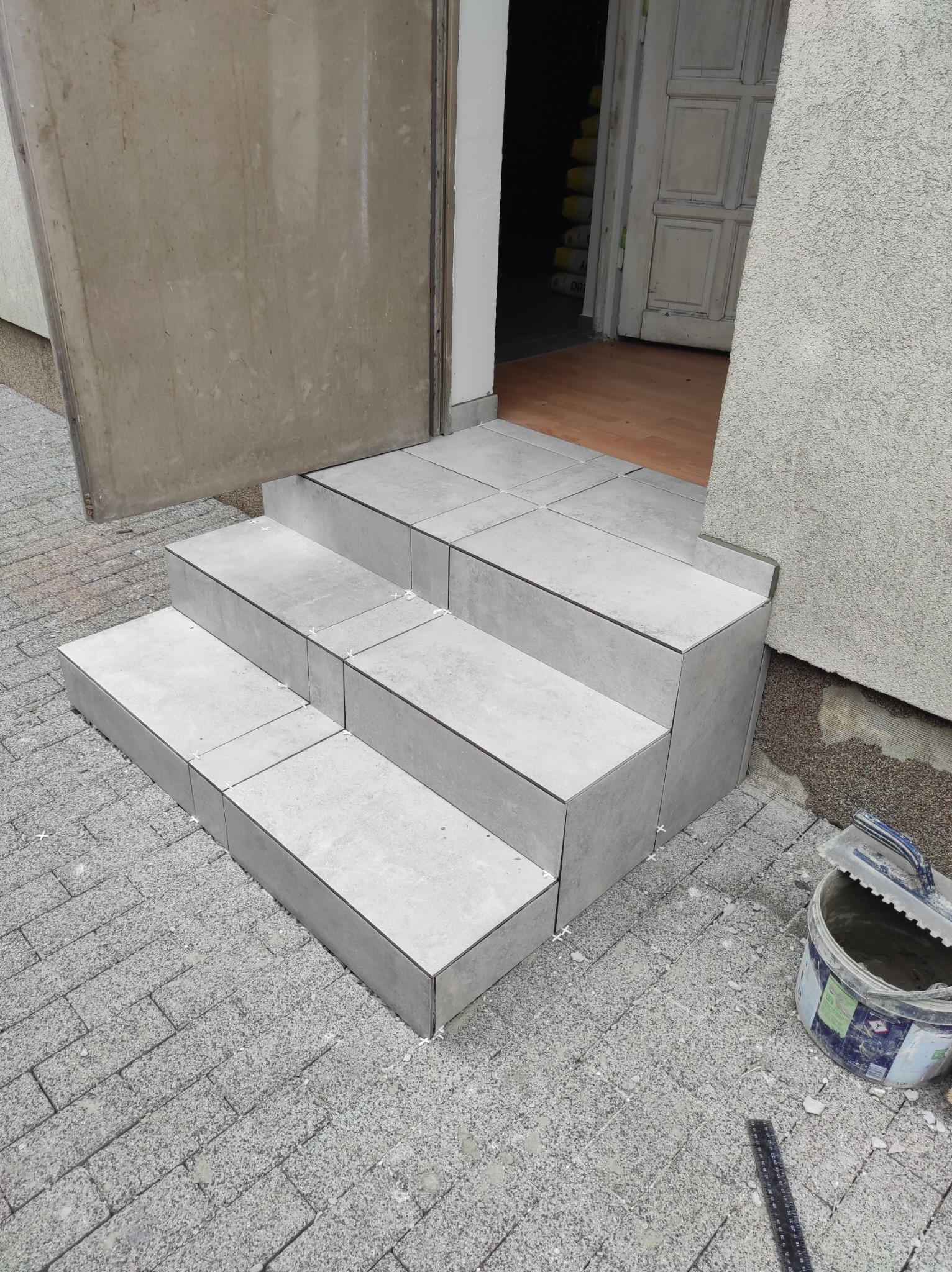 Wykończone schody wejściowe do budynku, wyłożone szarymi płytkami ceramicznymi, widoczne krzyżyki dystansowe i wiadro z zaprawą.