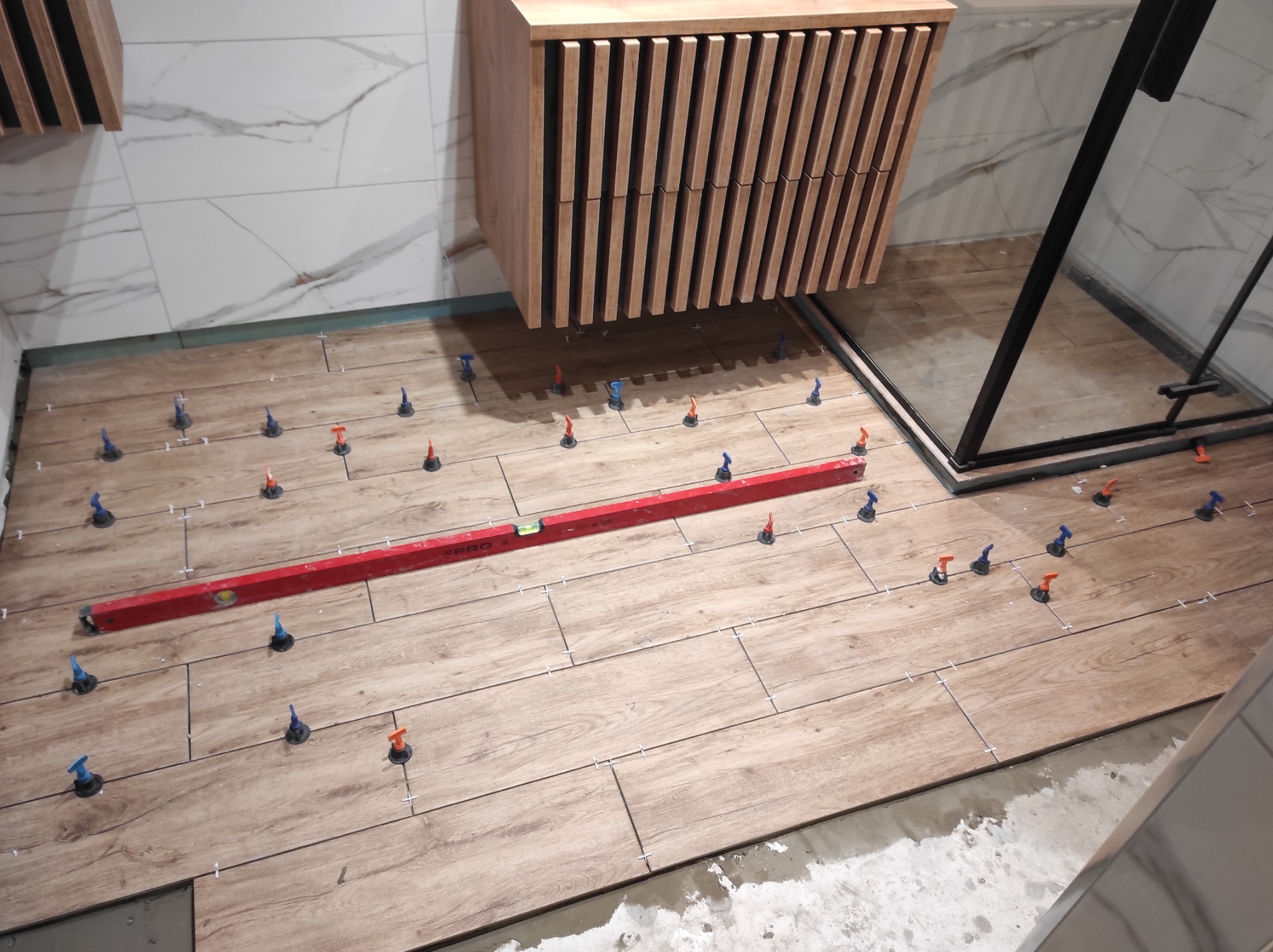 Układanie drewnopodobnych płytek podłogowych w łazience z użyciem poziomicy i systemu poziomowania, widoczna szafka pod umywalkę z lamelami i kabina prysznicowa.