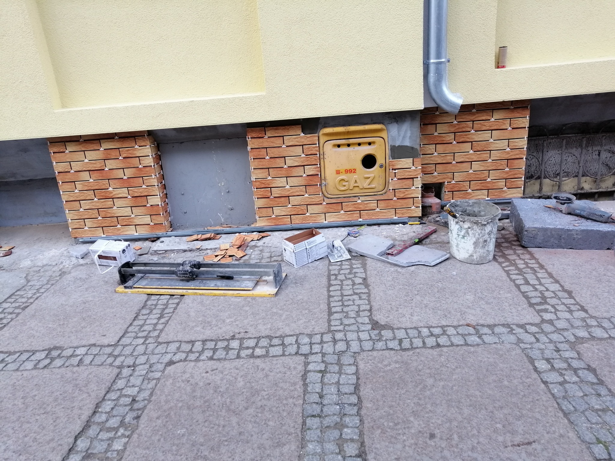 Fragment elewacji budynku z częściowo ułożoną cegłą klinkierową, widoczna skrzynka gazowa, narzędzia glazurnicze, wiadro i resztki płytek na kostce brukowej.