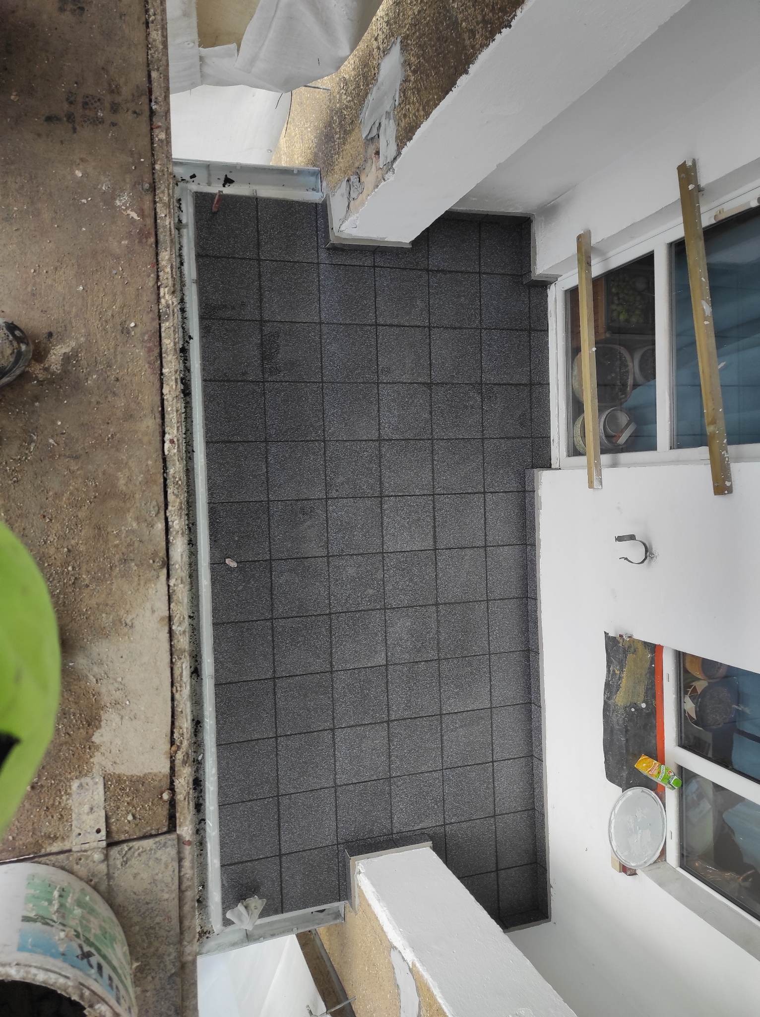 Widok z góry na balkon z szarą, kwadratową podłogą z płytek, z widocznymi elementami remontowymi: częściowo odrapana ściana, okno zabezpieczone taśmą malarską, wiadro z farbą w lewym dolnym rogu.