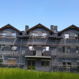 Trzysegmentowy budynek w trakcie prac elewacyjnych, pokryty szarym materiałem izolacyjnym, otoczony rusztowaniami, z widoczną drabiną i wysoką trawą na pierwszym planie.