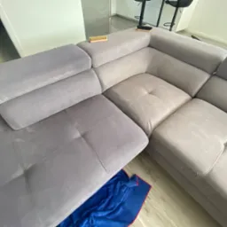 Fragment szarej, modułowej kanapy narożnej z widocznymi śladami czyszczenia i dwiema szczotkami na g&oacute;rze, z niebieską szmatką pod spodem, w jasnym, minimalistycznym wnętrzu.