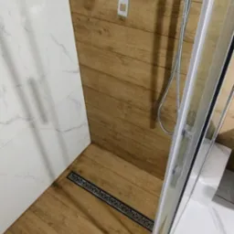 Nowoczesna kabina prysznicowa z odpływem liniowym w podłodze wyłożonej płytkami imitującymi drewno, bateria podtynkowa i deszczownica.