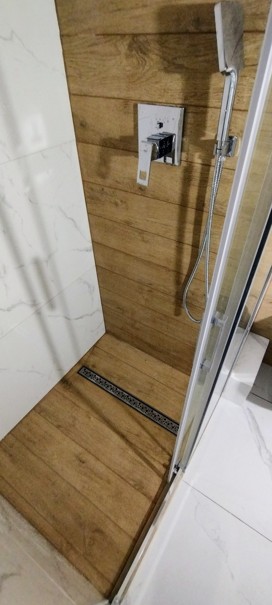 Nowoczesna kabina prysznicowa z odpływem liniowym w podłodze wyłożonej płytkami imitującymi drewno, bateria podtynkowa i deszczownica.