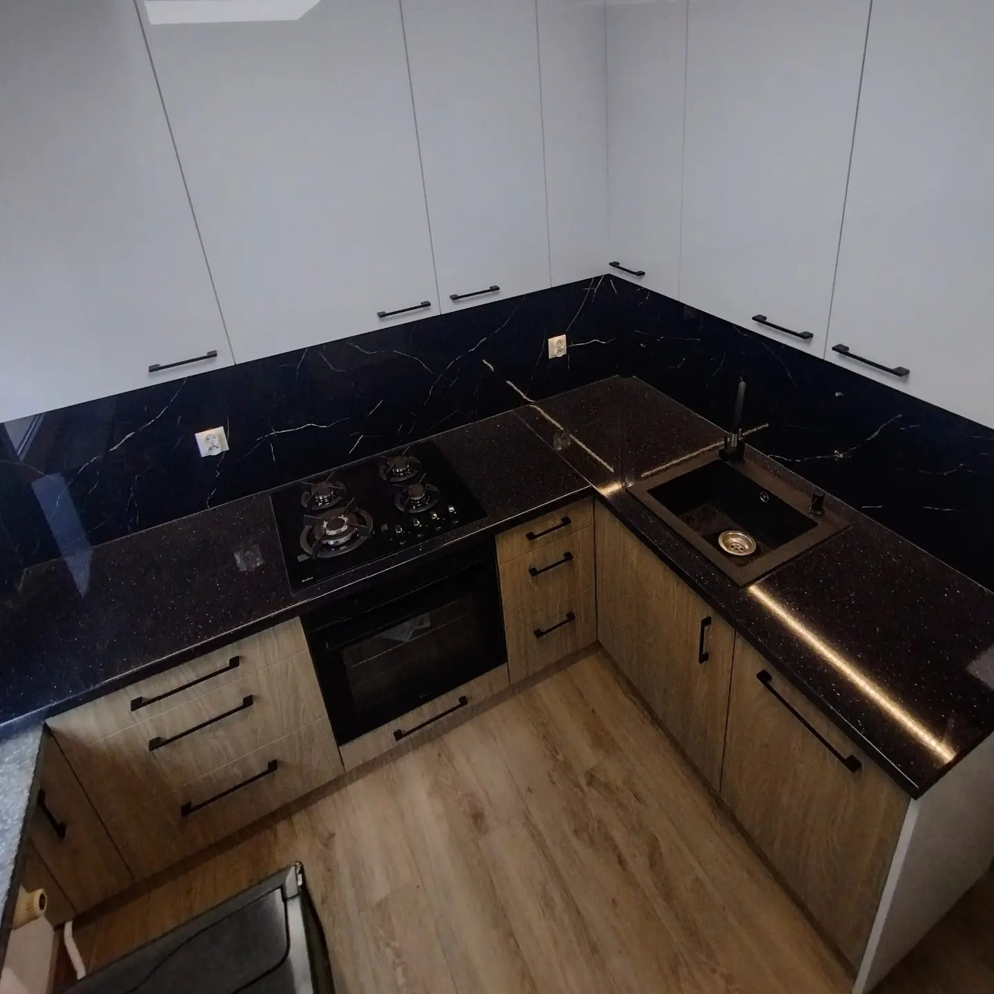 Nowoczesna kuchnia z białymi szafkami górnymi i drewnopodobnymi dolnymi, czarny blat z wbudowaną płytą grzewczą i zlewozmywakiem, oświetlenie LED pod blatem, czarne uchwyty, widok z góry.