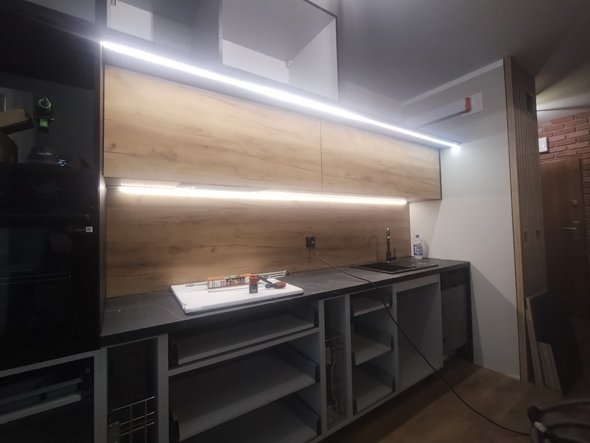 Montaż górnych szafek kuchennych z drewnianym fornirem i oświetleniem LED, widoczne narzędzia i materiały montażowe na blacie.