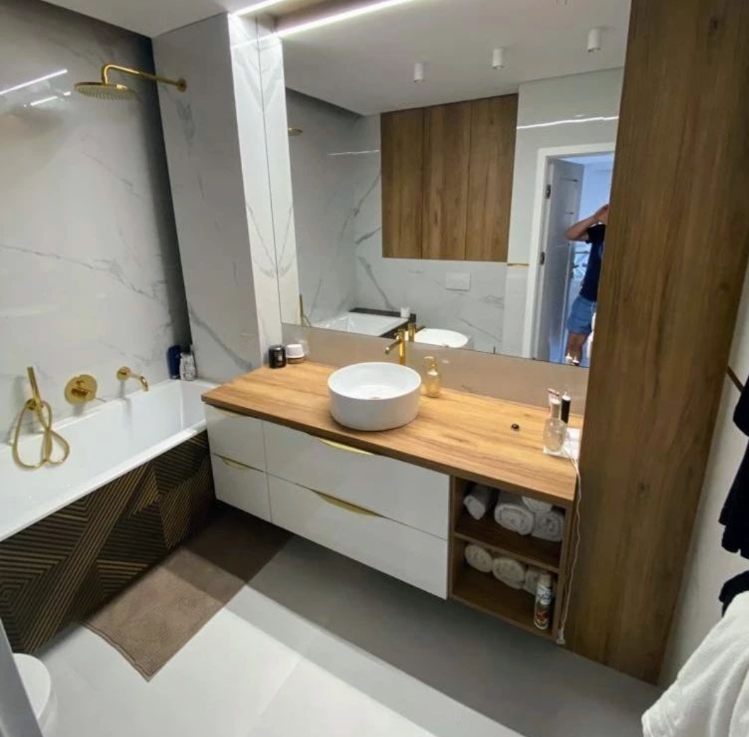 Nowoczesna łazienka z marmurowymi płytkami, złotą armaturą i drewnianym blatem pod umywalką, odbicie osoby w lustrze.