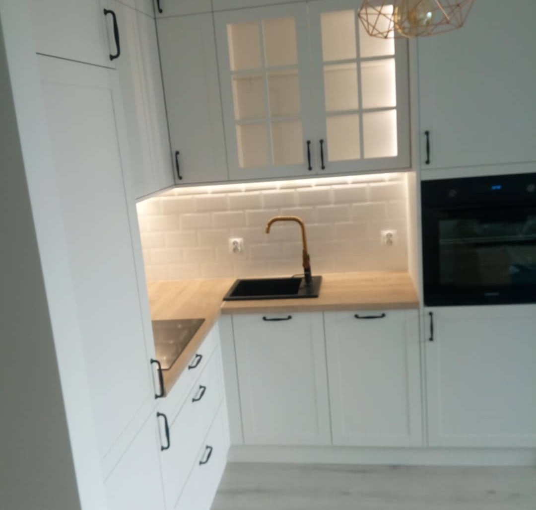 Jasna kuchnia z białymi szafkami, drewnianym blatem i złotą baterią, oświetlona listwą LED pod szafkami wiszącymi z przeszklonymi frontami, z widocznym czarnym zlewem.