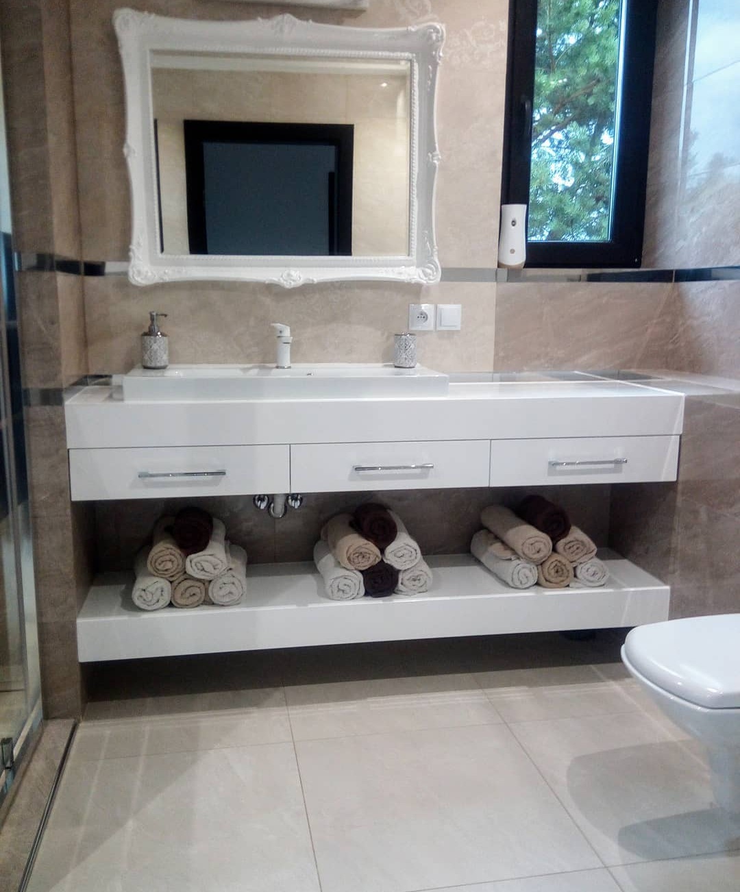 Jasna łazienka z umywalką w zabudowie, ozdobnym lustrem w białej ramie i rzędami zwiniętych ręczników na półce pod umywalką.