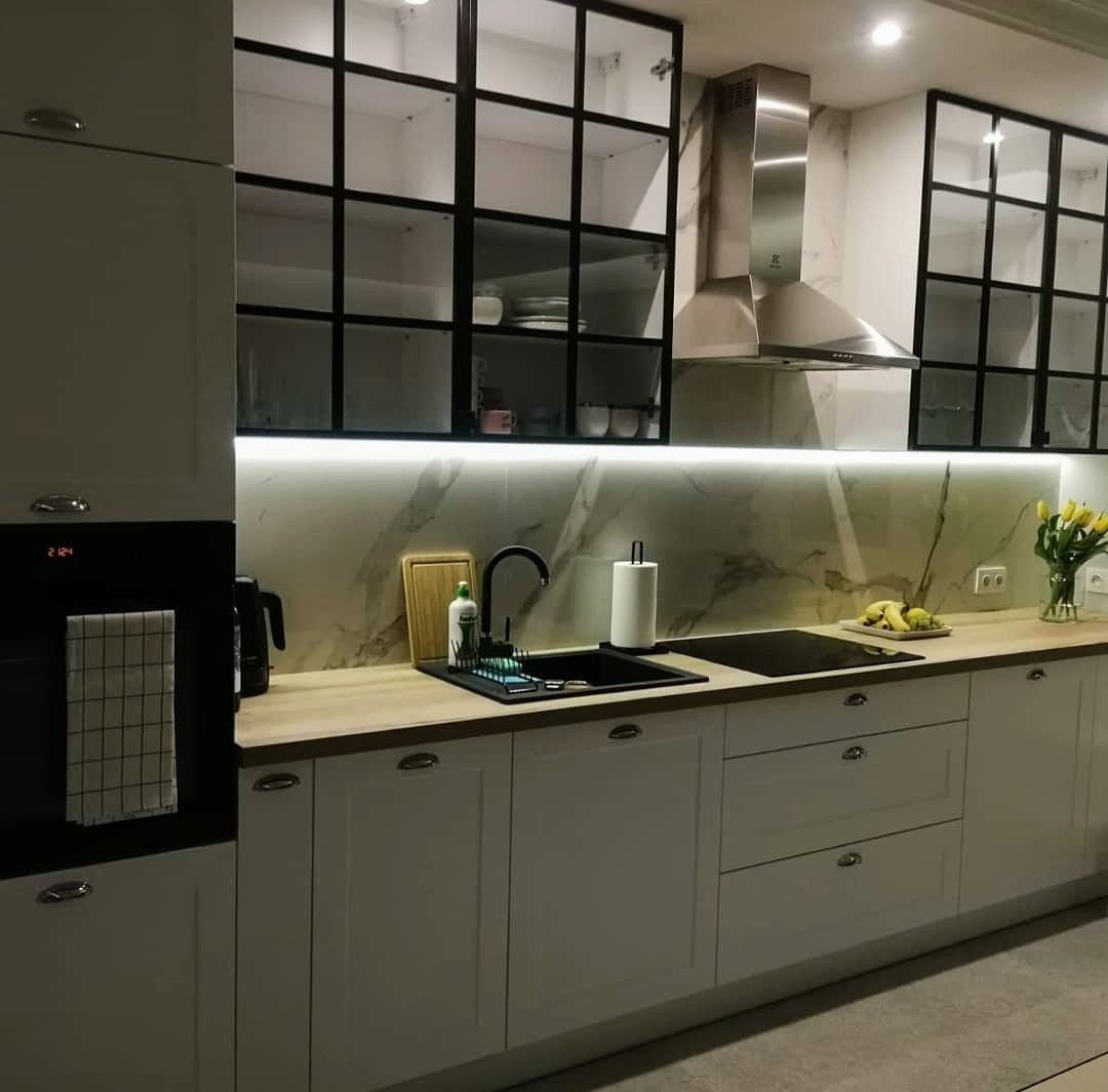 Jasna kuchnia z białymi szafkami, drewnianym blatem, czarnym zlewem i przeszklonymi szafkami wiszącymi w stylu industrialnym.