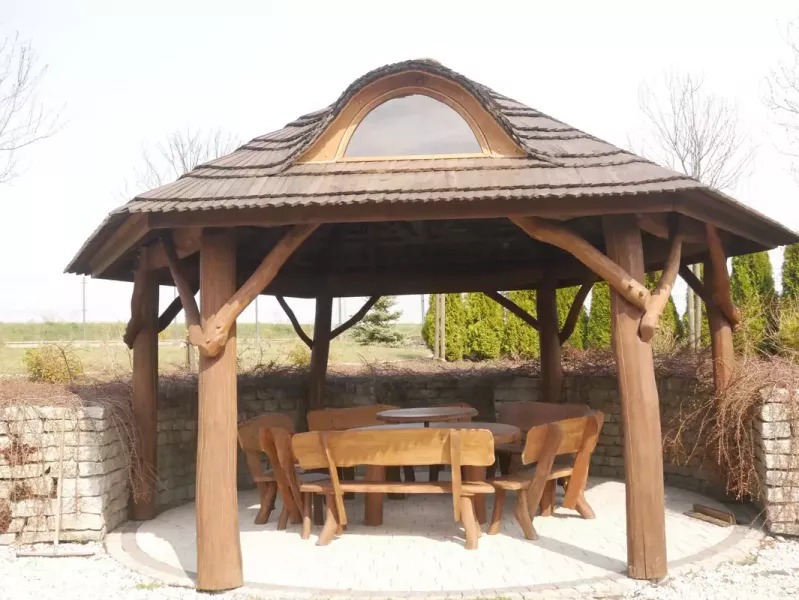 Drewniana altana ogrodowa z dachem krytym gontem, okrągłym oknem i meblami z litego drewna, otoczona kamiennym murkiem i jasną nawierzchnią.