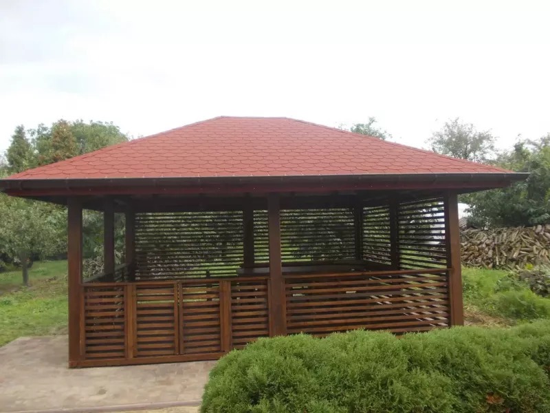 Zadaszona altana ogrodowa z brązowego drewna i czerwoną dachówką bitumiczną, otoczona zielenią i stosem drewna w tle.