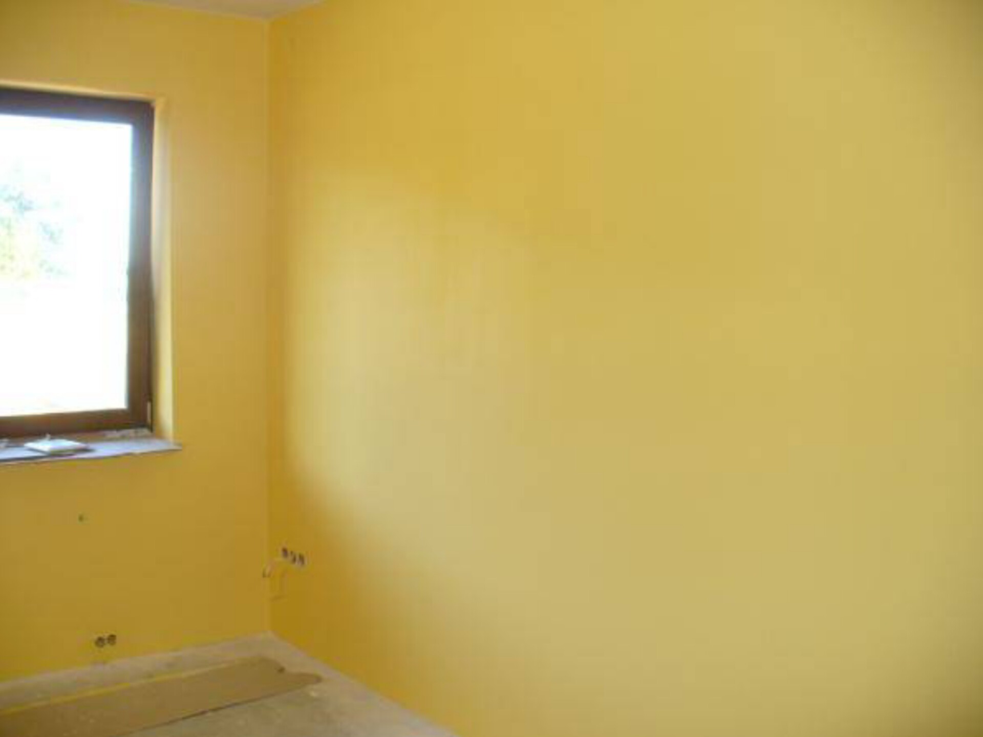 Pomalowane na żółto wnętrze pokoju z oknem, widoczne gniazdka elektryczne i fragment podłogi przed wykończeniem.