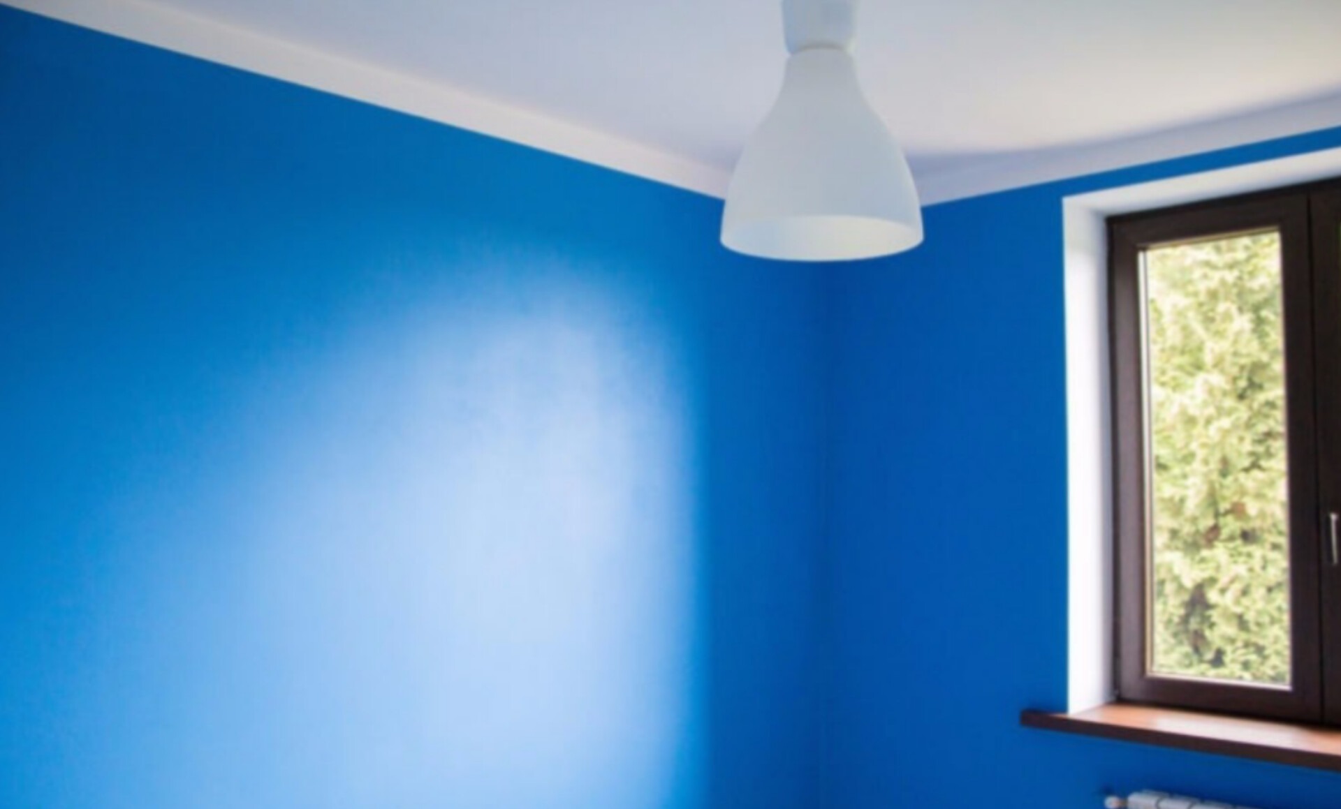 Pomieszczenie z intensywnie niebieskimi ścianami, białym sufitem z listwą, wiszącą lampą i oknem z brązową ramą, przez które widać zieleń drzew.