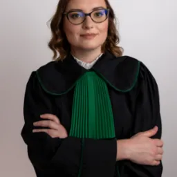 Portret kobiety w todze z zielonym żabotem i okularach, z założonymi rękami na piersi, na jasnym tle.