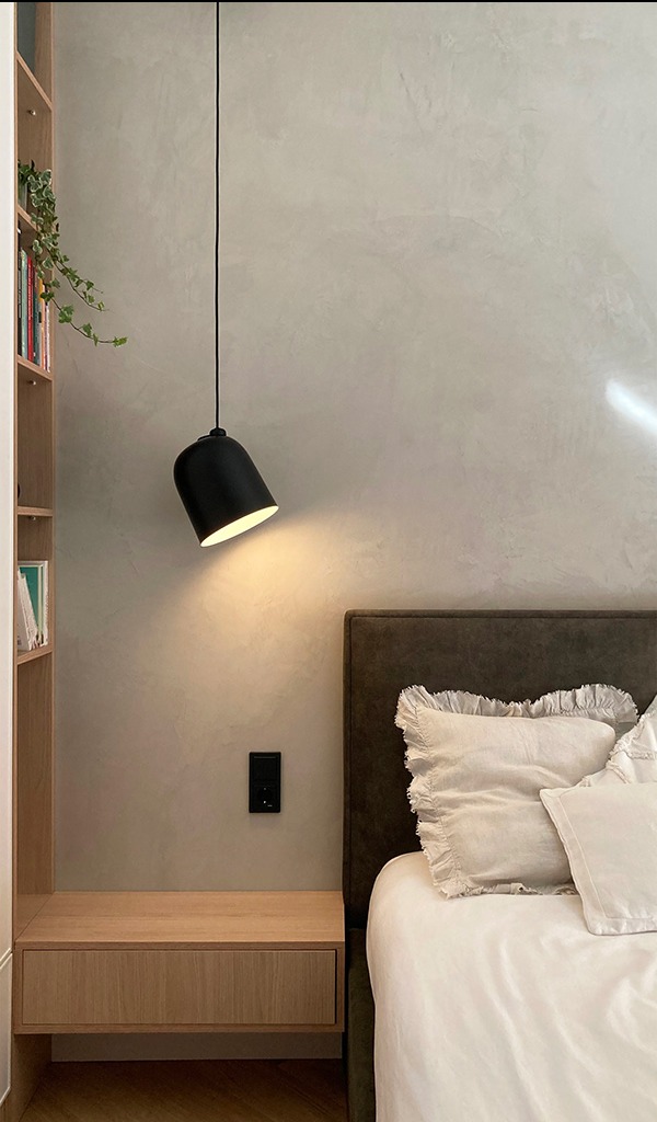 Minimalistyczna sypialnia z beżową ścianą o fakturze betonu, czarną lampą wiszącą nad drewnianą szafką nocną i łóżkiem z poduszkami z falbankami.