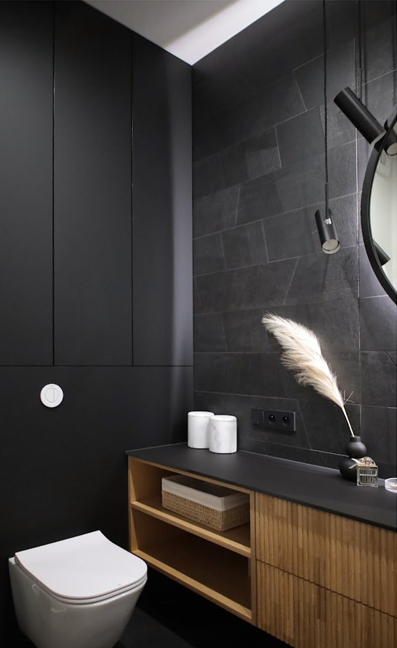 Nowoczesna, monochromatyczna łazienka z czarnymi płytkami imitującymi kamień, wiszącą toaletą i szafką z drewnianymi lamelami oraz dekoracyjnym piórem w wazonie.