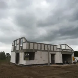 Surowy stan dwukondygnacyjnego domu z białych bloczków, widoczne otwory okienne i drzwiowe, szare nadproża, ziemisty teren wokół, pochmurne niebo w tle.