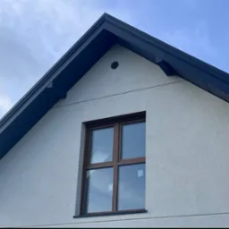 Fragment elewacji domu z ciemnym dachem i brązowym oknem, widoczny otw&oacute;r wentylacyjny na szczycie ściany.