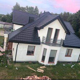 Lilek Usługi Dekarskie - Nowoczesny dom jednorodzinny z nowym, czarnym dachem z blachodachówki, widoczne rusztowanie i przygotowane drewno opałowe na trawniku.