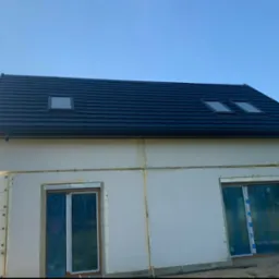 Dom w trakcie budowy z nowym, czarnym dachem z trzema oknami dachowymi i elewacją w trakcie ocieplania, okna zabezpieczone folią.