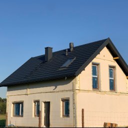 Lilek Usługi Dekarskie - Dom jednorodzinny w trakcie ocieplania elewacji i układania ciemnej dachówki, widoczne kominy i okno dachowe, okna zabezpieczone folią, budynek otoczony siatką ogrodzeniową.