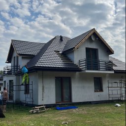 Lilek Usługi Dekarskie - Dekarz w trakcie wymiany pokrycia dachu z czarnej dachówki na dwupiętrowym domu z balkonem, widoczne rusztowanie i staw z liliami wodnymi w ogrodzie.