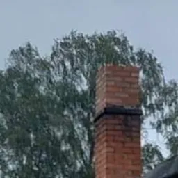 Murowany komin z cegły z widocznymi śladami okopcenia, wystający ponad fragment dachu pokrytego papą, na tle zielonych drzew i pochmurnego nieba.