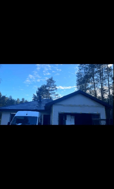 Dom z nowym, ciemnym dachem i częściowo ocieploną elewacją, zaparkowany biały bus widoczny z przodu, drzewa i błękitne niebo w tle.